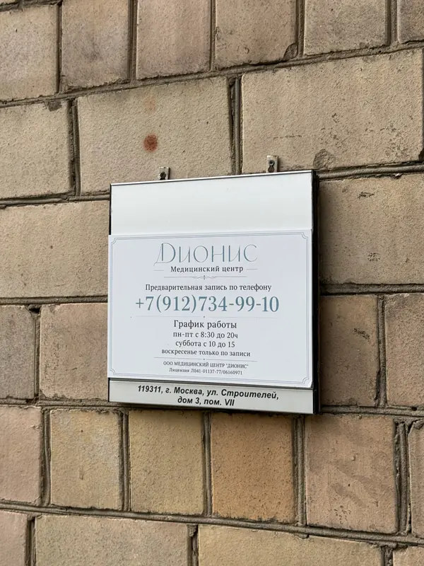 Вывеска медицинского центра «Дионис» на стене у входа: телефон и график работы